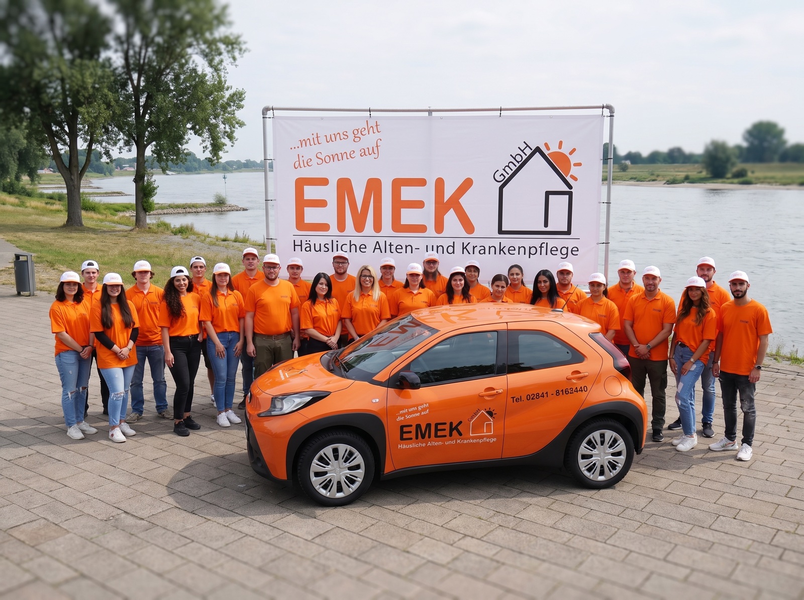 Das EMEK Team in orangen Shirts vor dem Firmenbanner an der Rheinpromenade, davor das orange EMEK Dienstfahrzeug